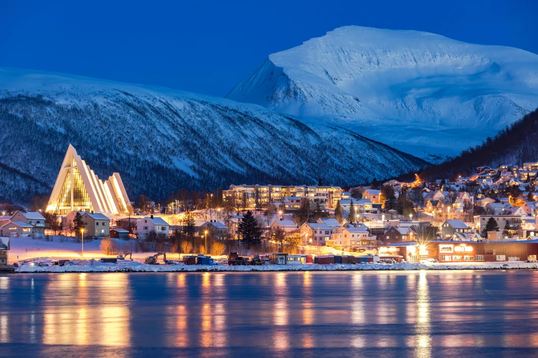 Atardecer sobre montañas nevadas con vistas a la Catedral del Ártico y casas tradicionales reflejadas en el fiordo helado, Tromso, Noruega